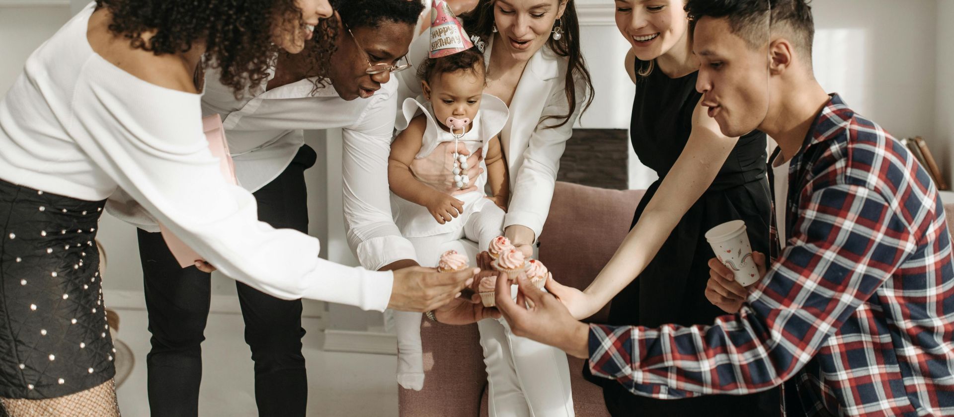 A diverse group celebrating a child's birthday indoors with cupcakes and party hats.